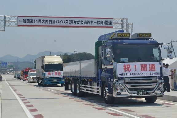 開通を祝う横断幕をつけてバイパスを走行する車両の写真