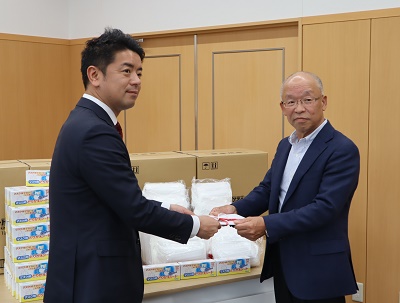 机に積まれたたくさんのマスクの前で寄贈品の目録を受け取っている様子の上村市長の写真