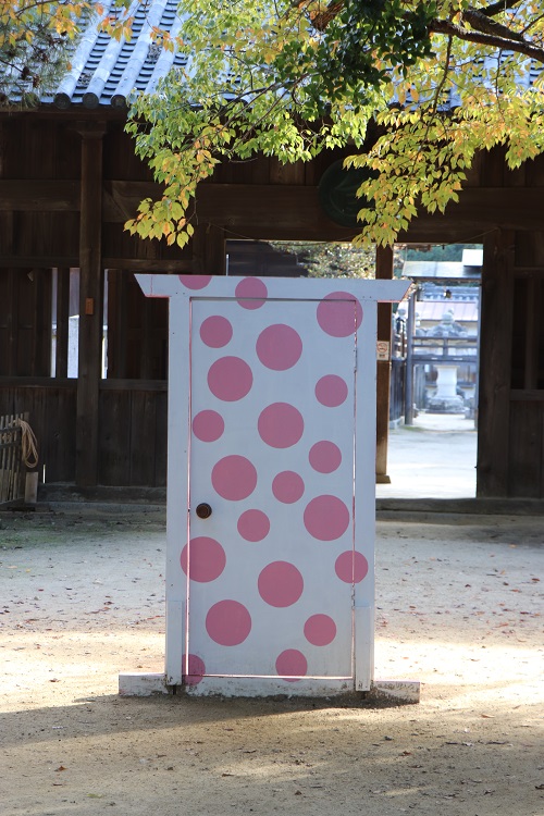 瓦屋根の木造の門の前に置かれた白地にピンクの水玉模様の扉の写真