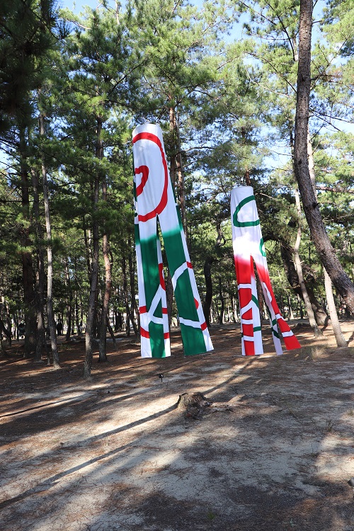 白い布のようなものに赤や緑で模様が描かれている造形物が木と木とあいだに吊るされている写真