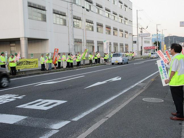 蛍光色のベストを身に着けた多くの関係者らが交通安全のプラカードを手に道路沿いに並んでいる様子の写真