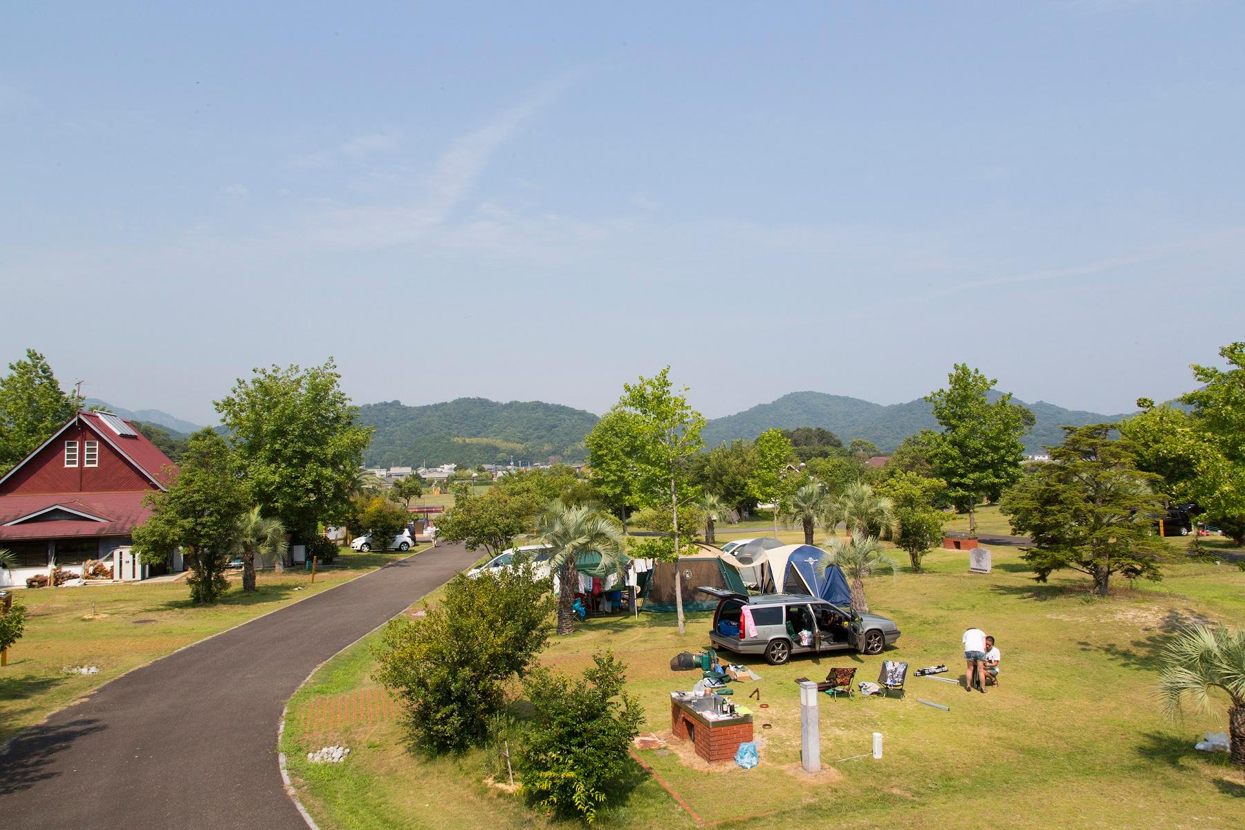 青空の下、緑の木々ときれいな芝生の大池オートキャンプ場に家族でキャンプの準備をしている様子の写真